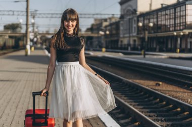 Beyaz etekli, kırmızı valizli, trenini bekleyen ve tren istasyonunda yürüyen esmer bir kadın yolcu.