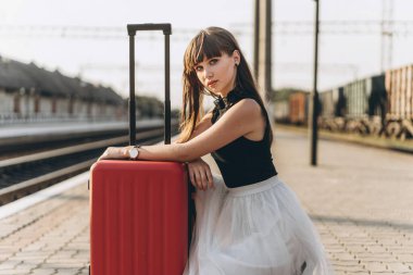 Beyaz etekli kırmızı valizli esmer kadın tren istasyonunda tren bekliyor.