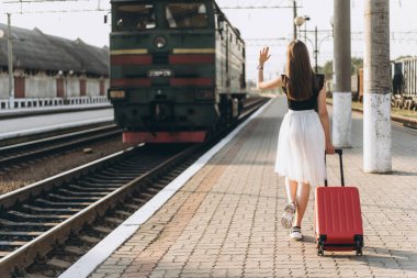 Beyaz etekli, kırmızı valizli, trenini bekleyen ve tren istasyonundaki trene el sallayan esmer bir kadın yolcu.