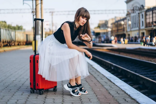 身穿白色短裙 头戴红色行李箱的女性黑发旅行者在候车站等火车 并在手表上查看时间 — 图库照片