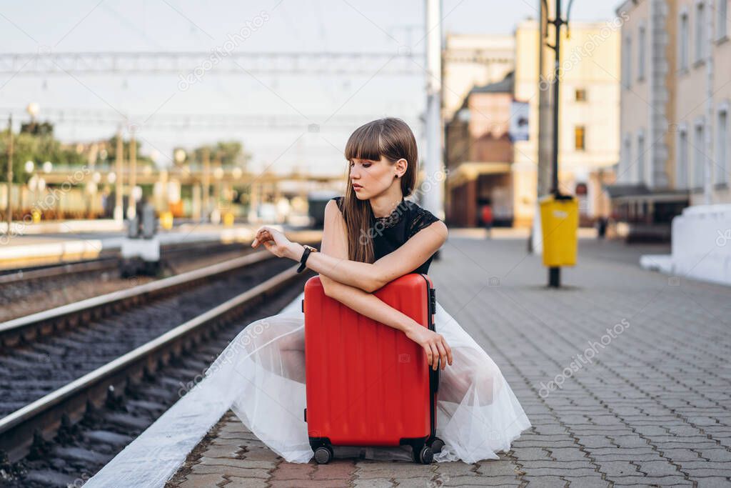 Female brunette traveler with red suitcase in white skirt waiting for a train on raiway station and checking time on her wristwatch