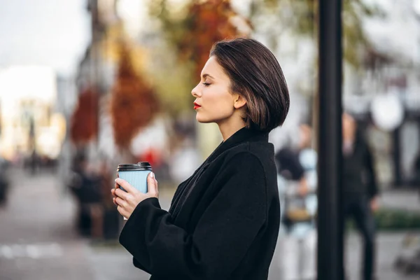 一个留着短发和红唇的年轻女人 手里拿着一杯咖啡 在城市的街道上漫步 周围都是人和车 — 图库照片