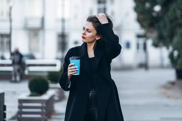一个红唇短发的年轻女人 身穿黑色衣服 摆姿势 手里拿着一杯咖啡 模糊的城市背景后面 — 图库照片
