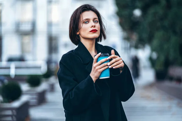 一个留着短发 身穿黑色衣服 摆姿势 手里拿着一杯咖啡的年轻女人 模糊的城市背景后面 — 图库照片