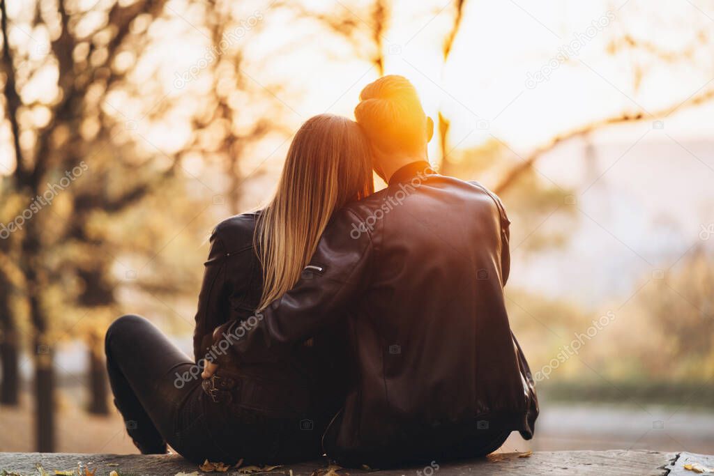 A romantic couple sitting on a bench hugging and watching the sunset in the park. Love story. Back view.