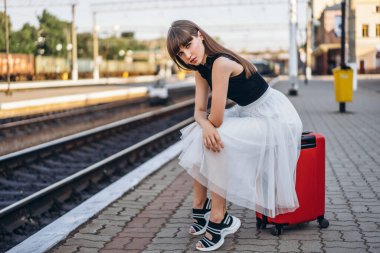 Beyaz etekli kırmızı valizli esmer kadın tren istasyonunda tren bekliyor.