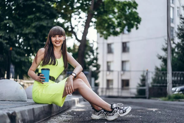 かなり長い髪ブルネット女の子で緑の光のドレスともに青紙カップの飲料座っている屋外で街の路上 — ストック写真