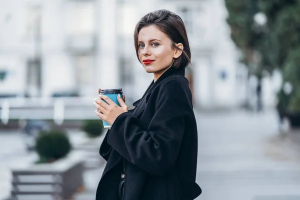 一个红唇短发的年轻女人 身穿黑色衣服 摆姿势 手里拿着一杯咖啡 模糊的城市背景后面 — 图库照片