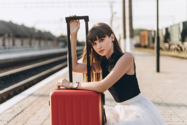 Beyaz etekli kırmızı valizli esmer kadın tren istasyonunda tren bekliyor.