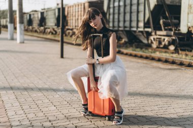 Beyaz etekli kırmızı valizli esmer kadın tren istasyonunda tren bekliyor.