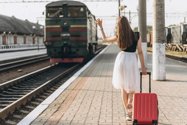 Beyaz etekli, kırmızı valizli, trenini bekleyen ve tren istasyonundaki trene el sallayan esmer bir kadın yolcu.