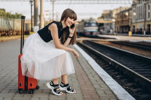身穿白色短裙 头戴红色行李箱的黑发女旅行者在火车站等火车 并使用手机 — 图库照片