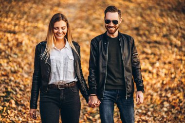 Güneşli bir günde sonbahar parkında yürüyen güzel genç bir çift. Sarılırlar, gülümserler, yürürken eğlenirler..