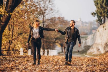 Sonbahar parkında gezen mutlu bir çiftin tam boy portresi. Erkek ve kadın el ele tutuşuyor.