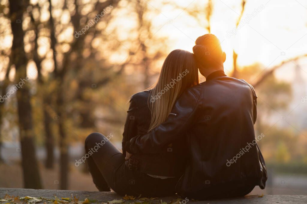 A romantic couple sitting on a bench hugging and watching the sunset in the park. Love story. Back view.