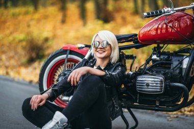 Güneş gözlüklü güzel sarışın motorcu kız ormanda kırmızı motosikletin yanında oturuyor. Renkli, bulanık bir geçmişi var..