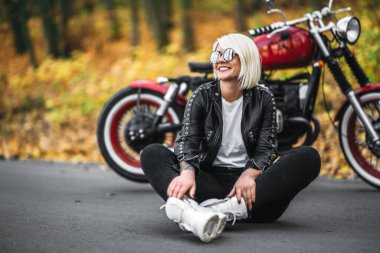 Güneş gözlüklü güzel sarışın motorcu kız ormanda kırmızı motosikletin yanında oturuyor. Renkli, bulanık bir geçmişi var..