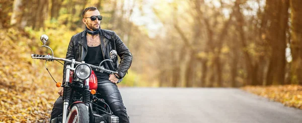 青白い背景を持つ森の道路上のオートバイに座ってサングラスや革のジャケットでひげを剃った残忍な男 — ストック写真