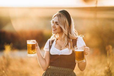 Geleneksel festival elbisesi içinde oldukça mutlu bir sarışındı. Arka planda bulanık iki kupa bira tutuyordu. Ekim Festivali, Aziz Patrick Günü, uluslararası bira günü konsepti.