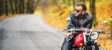 Güneş gözlüklü, deri ceketli, sakallı, vahşi bir adam ormanda bir motosikletin üzerinde oturuyor..