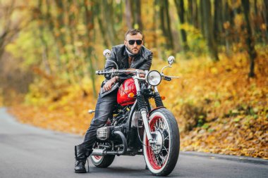 Güneş gözlüklü, deri ceketli, sakallı, vahşi bir adam ormanda bir motosikletin üzerinde oturuyor..