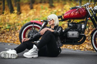 Güneş gözlüklü güzel sarışın motorcu kız ormanda kırmızı motosikletin yanında oturuyor. Renkli, bulanık bir geçmişi var..