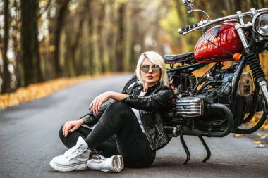 Güneş gözlüklü güzel sarışın motorcu kız ormanda kırmızı motosikletin yanında oturuyor. Renkli, bulanık bir geçmişi var..
