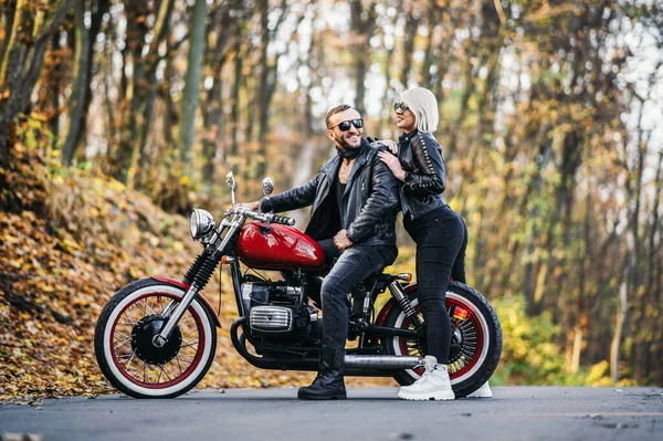 Ormandaki yolda, kırmızı motorsikletin yanında, renkli, mavi arka planda güzel bir çift. İlişki kavramı.