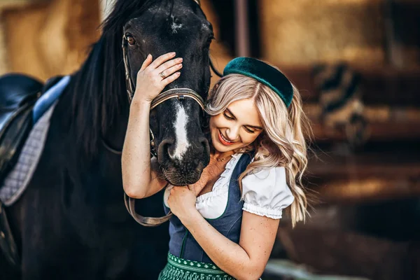 Geleneksel elbiseli güzel sarışın çiftlikteki büyük siyah atla ilgileniyor. Hayvanlar ve insanlar dostluk kavramı. 