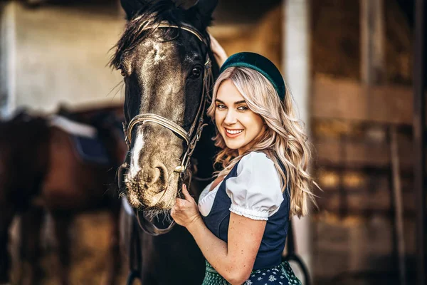 Geleneksel elbiseli güzel sarışın çiftlikteki büyük siyah atla ilgileniyor. Hayvanlar ve insanlar dostluk kavramı. 