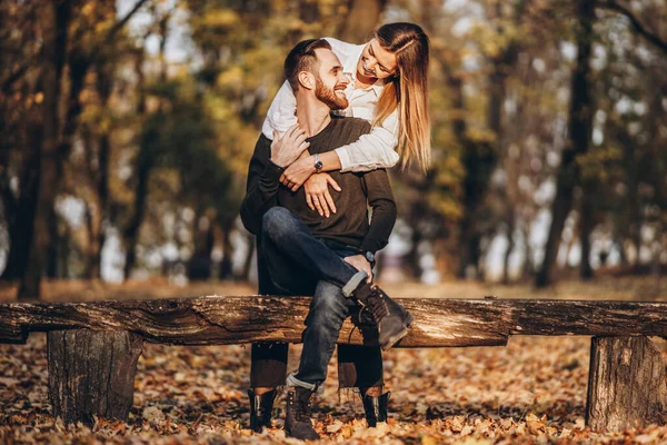 森の中の木製のベンチに座っている若い愛情のあるカップル 秋の木々の中で男と女が抱き合って笑っている — ストック写真