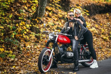 Ormandaki yolda, kırmızı motorsikletin yanında, renkli, mavi arka planda güzel bir çift. İlişki kavramı.