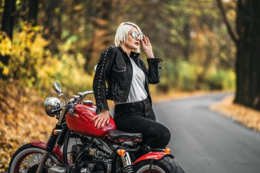 Kırmızı motosikletli, güneş gözlüklü güzel sarışın motosikletli kız. Ormanda yol kenarında. Renkli, bulanık bir geçmişi var..