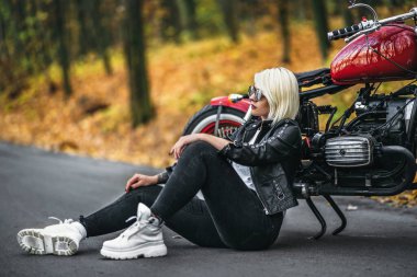 Güneş gözlüklü güzel sarışın motorcu kız ormanda kırmızı motosikletin yanında oturuyor. Renkli, bulanık bir geçmişi var..