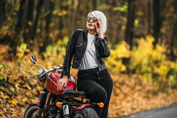 美しいですブロンドバイカー女の子でサングラスで赤いオートバイとともに道路上の森の中で色とりどりのぼやけた背景 — ストック写真