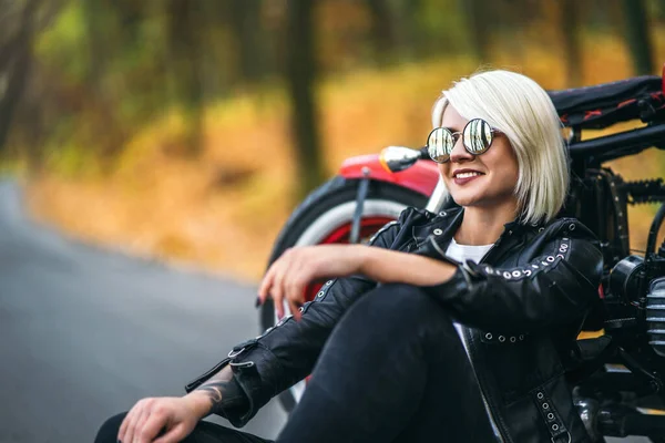 Güneş gözlüklü güzel sarışın motorcu kız ormanda kırmızı motosikletin yanında oturuyor. Renkli, bulanık bir geçmişi var..