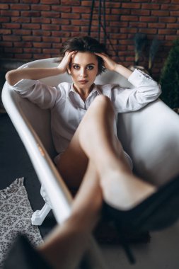 Güzel bir esmerin portresi, boş bir banyoda uzanmış, bacakları havada, beyaz gömlek ve siyah ayakkabılar giymiş, kırmızı tuğlalı arka planda..