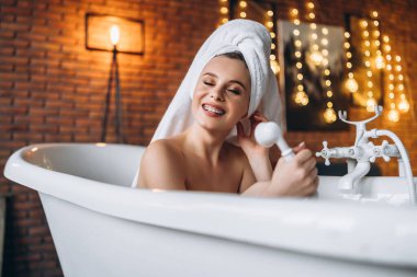 Stüdyoda poz veren güzel bir genç kadın. Kafasında havluyla beyaz bir banyoda yatan bir kadın. Arka planda sarı çelenkler olan kırmızı tuğladan bir duvar..