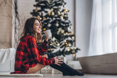 Güzel esmer kız kırmızı gömlekli ve sıcak çoraplı bir şekilde yerde oturuyor ve arkasında bir fincan kahve ve Noel ağacı var.