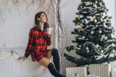 Yatakta güzel esmer kız kırmızı gömlekli ve sıcak çoraplı bir fincan kahve ve Noel ışıkları arkasında.