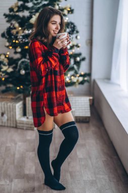 Güzel esmer kız pencerenin yanında duruyor, kırmızı gömlek ve sıcak çoraplar giyiyor ve arkasında bir fincan kahve ve Noel ışıkları var.