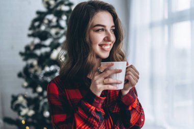 Güzel esmer kız pencerenin yanında duruyor, kırmızı gömlek ve sıcak çoraplar giyiyor ve arkasında bir fincan kahve ve Noel ışıkları var.