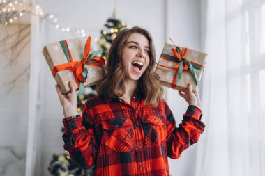 Noel, yeni yıl. Gömlekli ve çoraplı güzel bir kadın Noel hediyesi kutusuyla evde yürüyor, yılbaşı ağacı arkasında.