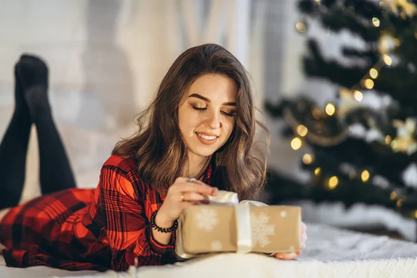 Noel, yeni yıl. Gömlekli ve çoraplı güzel bir kadın Noel hediyesi kutusuyla yatakta yatıyor, yeni yıl ağacı arkasında.