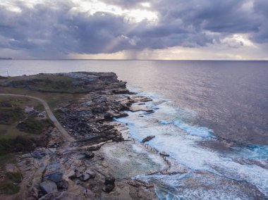Maroubra 'daki Mahon kaya havuzunun havadan görünüşü ve Avustralya' nın güzel kıyı şeridi. Yoğun bulutları delmeye çalışan güneş ışığıyla çarpıcı bir gökyüzü var.