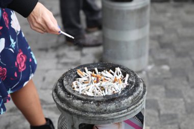 Cigarette in the hands of a woman next to an ashtray with a lot of cigarette butts in the city clipart