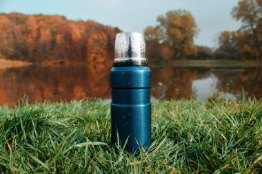 Yeşil çimlerde mavi elektrikli süpürge termosu, sonbahar ormanı ve göl arka planı. Yürüyüş, açık hava eğlencesi, turizm....