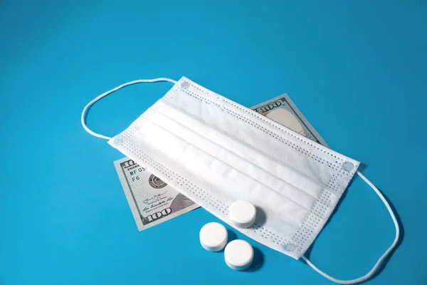 Pills and medical face mask covered one hundred dollar bill on blue background. Concept of economic crisis caused by coronavirus pandemic, expensive treatment and vaccine.