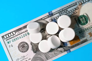 Mavi arka planda hapların altında yüz dolarlık banknot, yakın plan. Bedjamin Franklin ilacın etkisinden kurtuluyor. Artan uyuşturucu fiyatları, sağlık bütçesi, ilaç ve tedavi maliyetleri