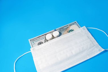Tıbbi yüz maskesi altında mavi arka planda 100 dolar, kopyalama alanı. Haplar Benjamin Franklin 'in önünde duruyor. Küresel durgunluk, covid-19, sağlık ve sağlık harcamaları.